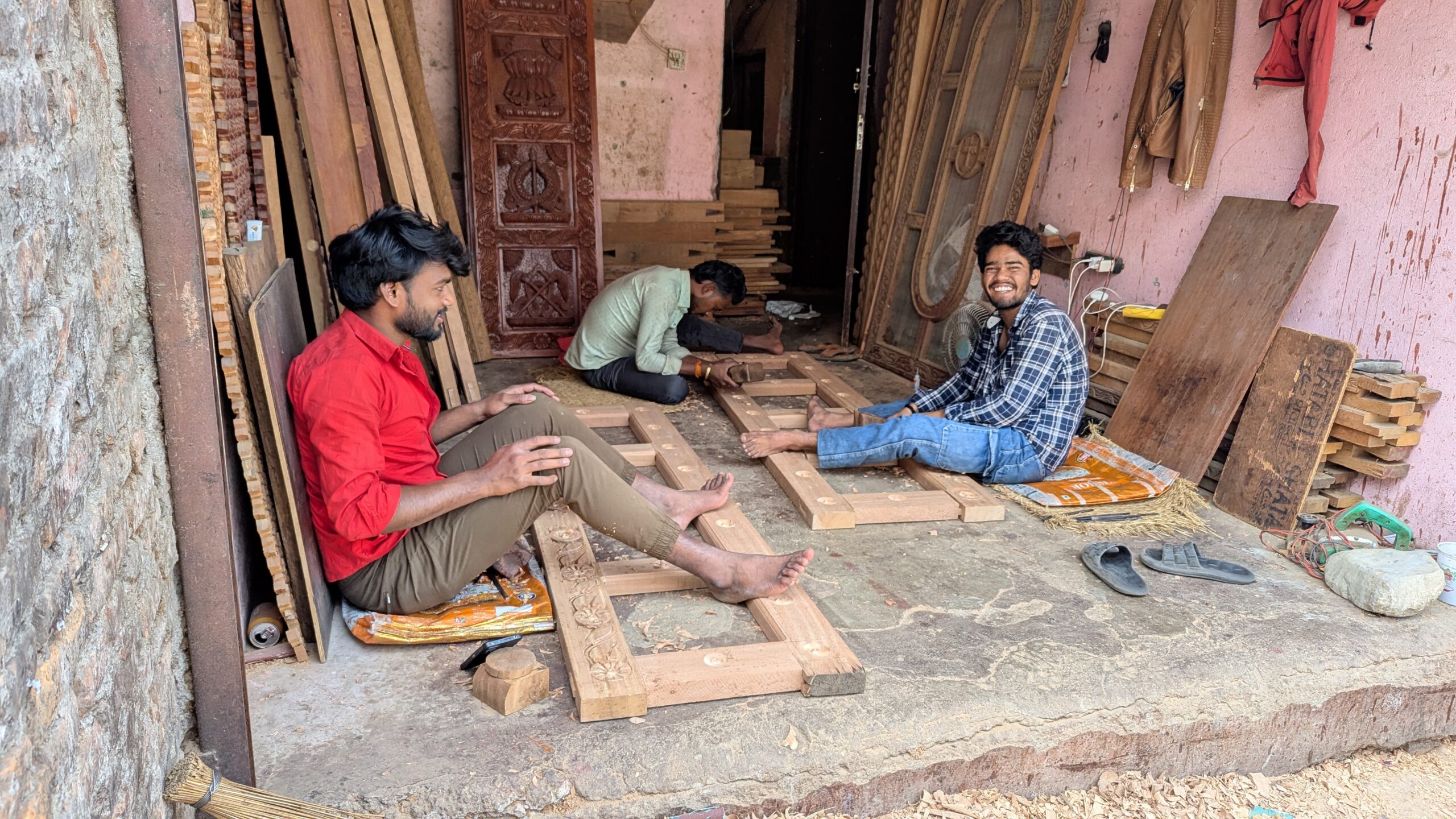 Three men sitting on the floor of an open workshop, hand chiseling wood.